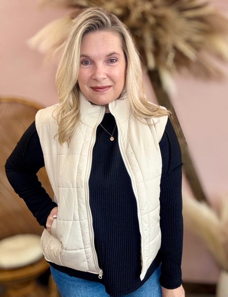 Woman wearing a cream quilted vest over a black top with a blurred indoor background
