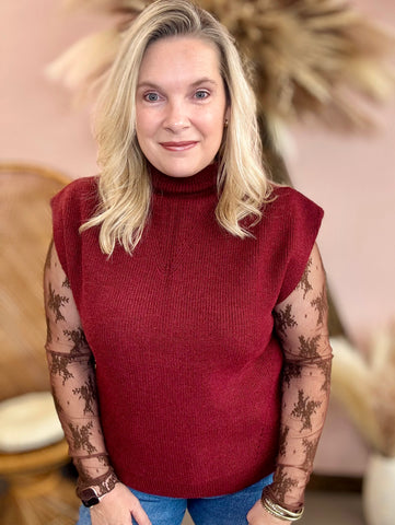 Woman wearing a red sleeveless sweater with floral arm coverings in a blurred indoor setting
