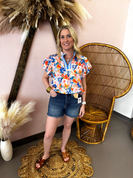 Woman in a floral shirt and denim shorts standing in a room with decorative elements.