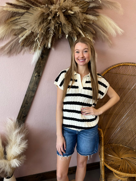 Young woman wearing a striped shirt and denim shorts standing in front of decorative plants and a wicker chair.