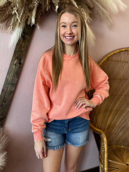 Woman wearing a coral sweatshirt and denim shorts standing in front of decorative elements.