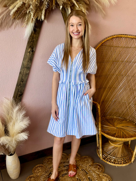 Woman in a blue and white striped dress standing in a decorative setting with pampas grass and a wicker chair.
