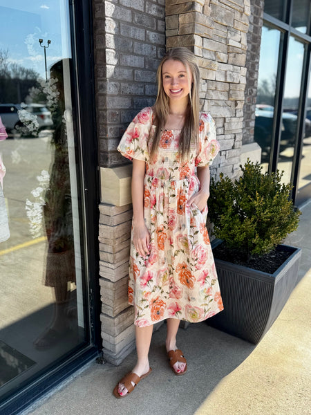 Woman in a floral dress standing outside a building with a stone wall and glass door.