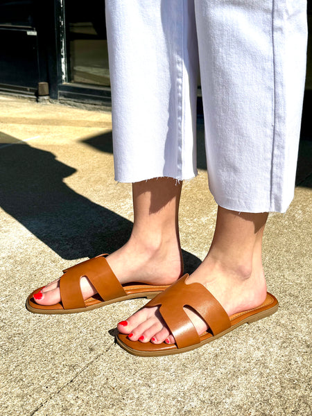 Brown sandals worn with white pants on a concrete surface.