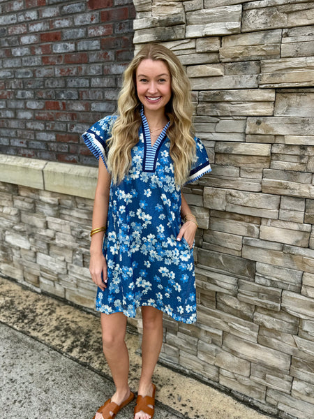 Woman in a blue floral dress standing against a stone wall.