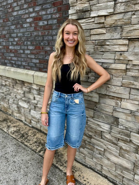 Woman in black top and blue denim shorts standing against a stone wall.