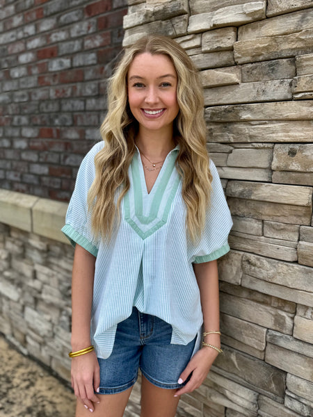 Woman wearing a light blue striped shirt and denim shorts standing against a stone wall.