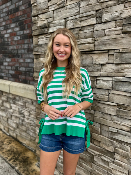Woman wearing a green and white striped shirt and denim shorts standing against a stone wall.