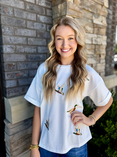 Woman wearing a white t-shirt with mallard duck designs standing in front of a brick wall.