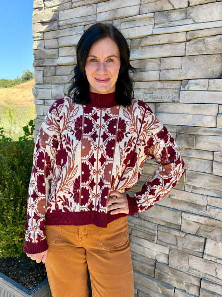 A woman wearing a burgundy and cream patterned knit sweater with a mock neck and brown pants, standing outdoors against a gray stone wall.