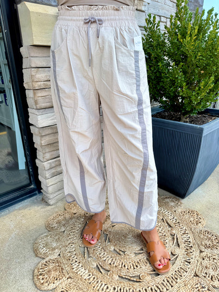 A person wearing loose, wide-leg linen pants in a light beige color with gray side stripes, standing on a woven doormat outdoors next to a potted plant.
