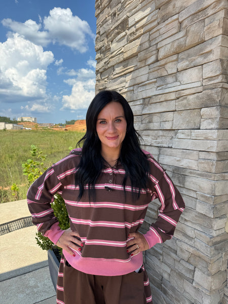 A woman with long black hair stands outdoors against a stone wall, wearing a brown and pink horizontally striped long-sleeve top and matching brown pants.