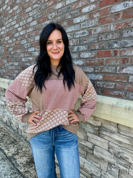 A woman with long black hair stands against a brick wall wearing a color-blocked long-sleeve top with beige, pink-and-white striped, and beige geometric patterned sections, paired with blue jeans.