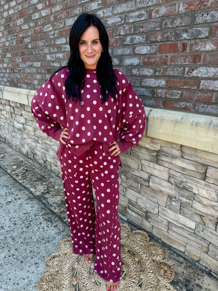 A woman modeling a matching two-piece lounge set consisting of a long-sleeve sweatshirt and wide-leg pants, both in a deep burgundy color with white polka dots, standing outdoors against a brick and stone wall.
