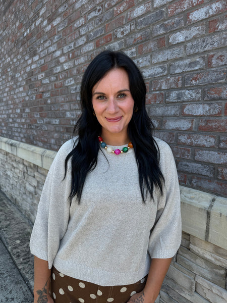 A woman with long black hair stands outdoors against a brick wall, wearing a light beige knit sweater and a brown polka-dot skirt, accessorized with a colorful beaded necklace.