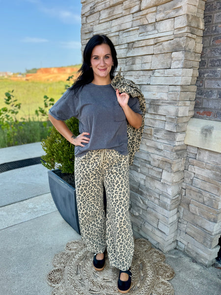 A woman stands outdoors against a stone pillar wearing a loose dark gray t-shirt, leopard-print wide-leg pants, and black flats, with a matching leopard-print cardigan draped over her shoulder.