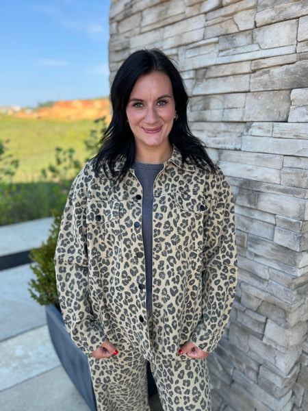 A woman wearing a beige and black leopard print jacket and matching pants, standing outdoors against a stone wall.