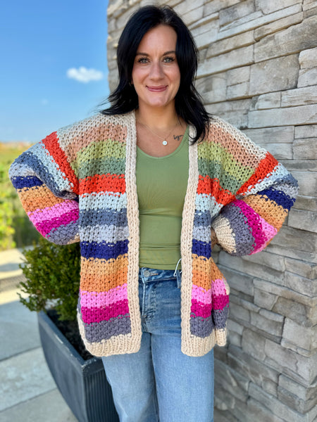 A woman wearing a chunky, open-front crochet cardigan with horizontal rainbow stripes in beige, orange, pink, purple, blue, green, and white, paired with a green tank top and blue jeans, standing outdoors against a stone wall.