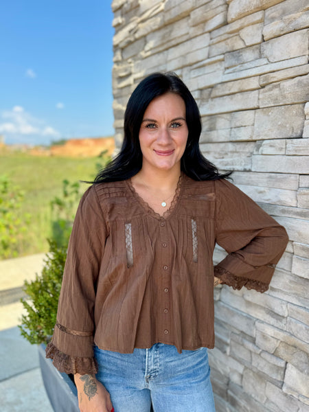 A woman with long black hair stands outdoors against a light stone wall, wearing a brown long-sleeve button-front blouse with lace trim and light blue jeans.