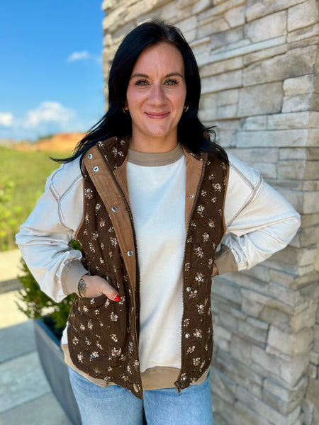 A woman wearing a brown floral-patterned zip-up vest over a cream sweatshirt and light blue jeans, standing outdoors against a stone wall.