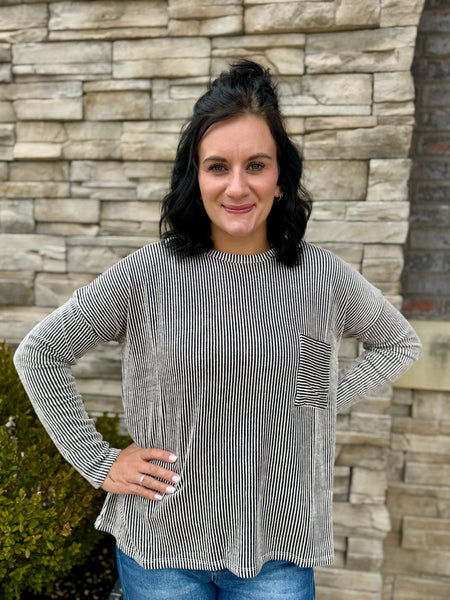 Black and white striped long-sleeve top with a front pocket, paired with medium-wash blue jeans, styled on a model against a stone wall.