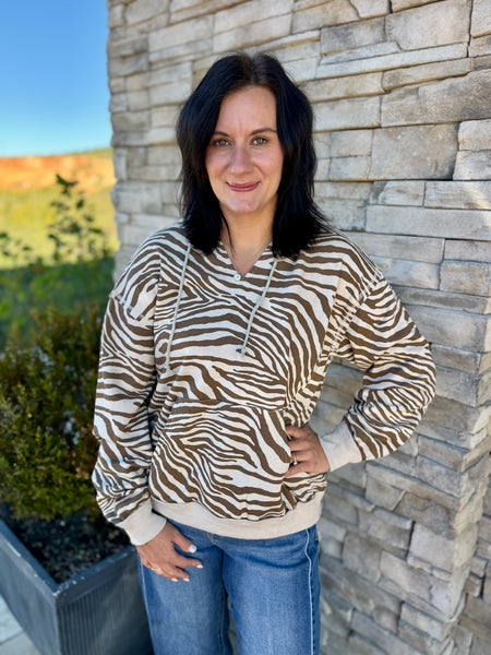 A woman wearing a brown and beige zebra-print hoodie stands outdoors against a stone wall, with blue jeans visible below.