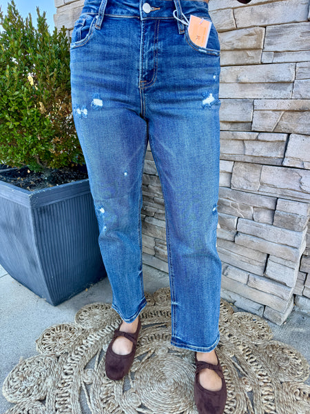 A person wearing medium-wash blue distressed jeans with small white bleach spots, paired with dark brown flat shoes, standing outdoors against a stone wall and planter.
