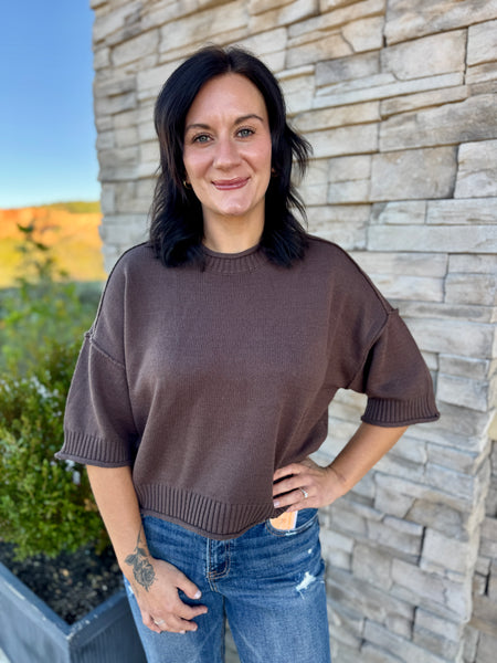 A woman with dark shoulder-length hair stands outdoors in front of a light stone wall, wearing a loose brown short-sleeve knit sweater and blue jeans.