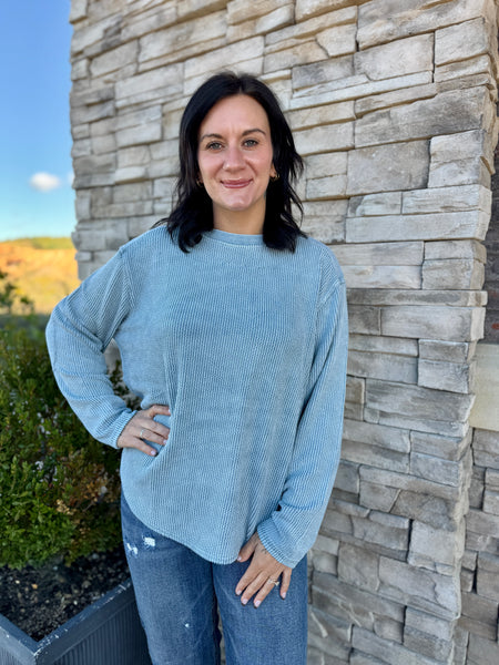 A woman wearing a light blue ribbed long-sleeve top and blue jeans, standing outdoors against a stone wall.