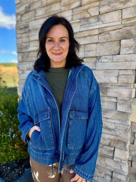 A woman wearing a blue denim jacket over an olive green shirt and brown pants, standing outdoors in front of a stone wall.