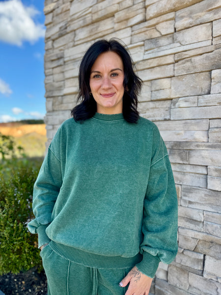 A woman with dark shoulder-length hair stands outdoors in front of a light stone wall, wearing an oversized muted green sweatshirt and matching green pants.
