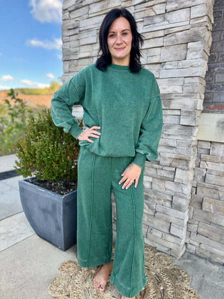 A woman modeling a matching green lounge set consisting of a cropped long-sleeve sweatshirt and wide-leg pants, standing outdoors in front of a stone wall.