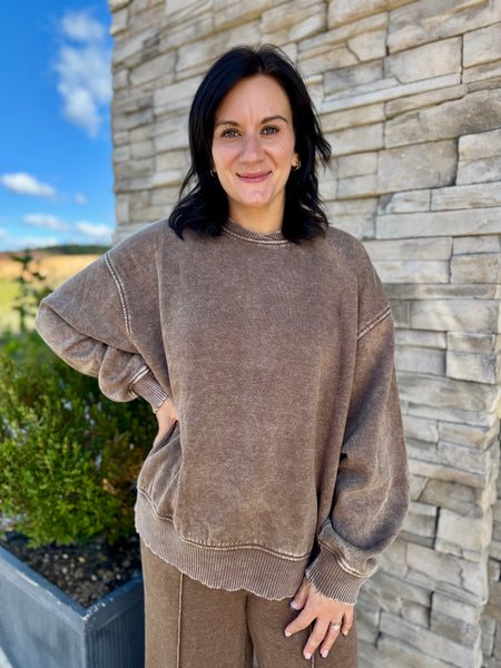 A woman wearing a brown acid-wash crewneck sweatshirt and matching sweatpants, standing outdoors in front of a stone wall.