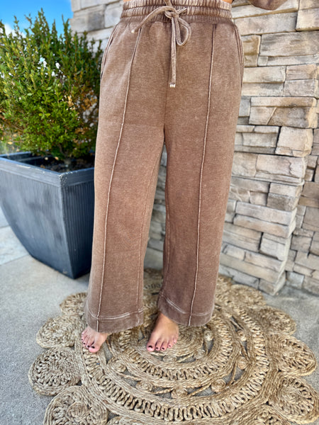 A person wearing wide-leg brown drawstring pants stands barefoot on a round woven rug in front of a stone wall and potted plant.