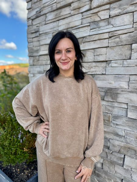 A woman with dark hair wearing a beige corduroy sweatshirt and matching sweatpants, standing outdoors against a gray stone wall.