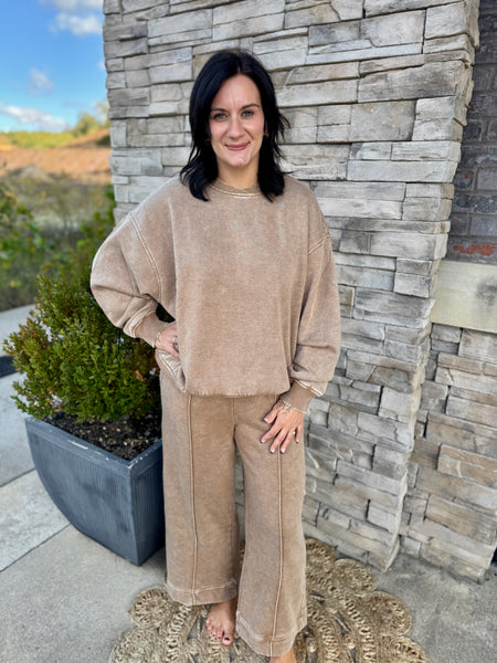 A woman wearing a matching beige lounge set consisting of a cropped long-sleeve sweatshirt and wide-leg pants, standing outdoors in front of a stone wall.