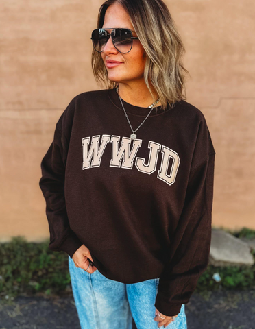 Woman wearing a brown sweatshirt with 'WWJD' text against a beige wall.
