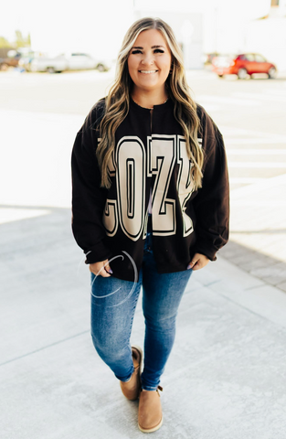 Woman wearing a brown sweatshirt with 'COZY' text, blue jeans, and brown shoes outdoors.